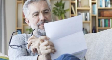  Man with grey hair and beard holding glasses sits on his couch at home reviewing Highmark Medicare Plans paperwork