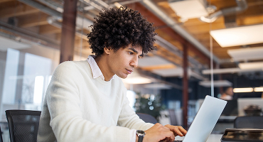  A young man types intently on his laptop in a bustling office space.