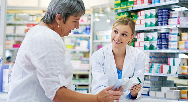 A pharmacist in a white lab coat smiling and discussing medication with an older female customer in a pharmacy.