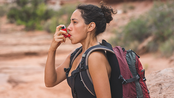 A person with a backpack stops on a rocky trail to use a red inhaler to help them breathe, with desert plants and red rock formations in the background.