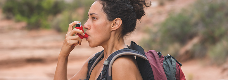 A person with a backpack stops on a rocky trail to use a red inhaler to help them breathe, with desert plants and red rock formations in the background.