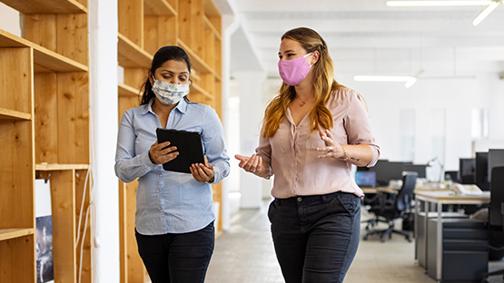 Two adults wearing face masks walk and talk in an office; one person holds a tablet.
