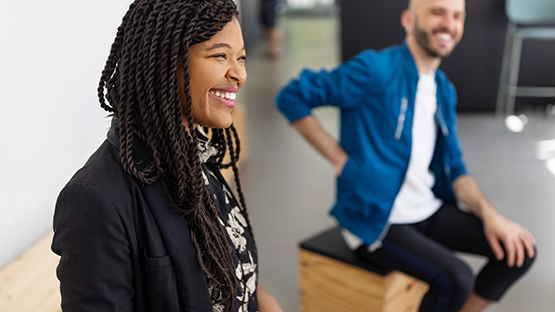 A person with long braids smiles while sitting, and another person sits behind them, smiling too.