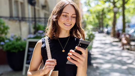A young woman is walking down a sunny street and looking at her phone with a smile on her face.
