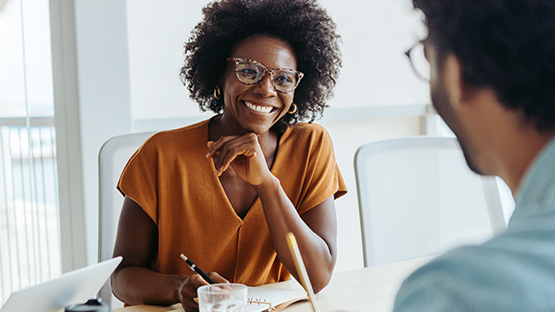A smiling person with curly hair and glasses, wearing a rust-colored shirt, sits at a table across from another person whose back is visible.