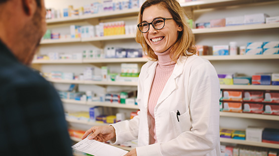 A smiling medical professional in a white lab coat and glasses hands a paper to a person with their back to the camera in a pharmacy.