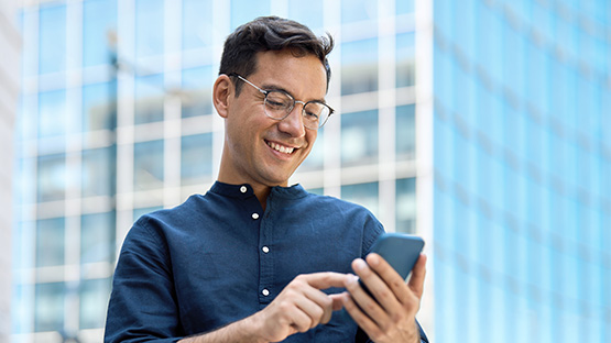 A smiling young man in glasses and a dark blue shirt is looking down at his phone and typing. A modern building with blue windows is blurred in the background.