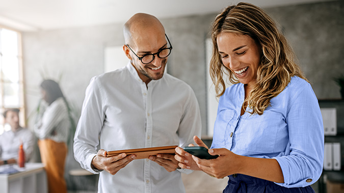A bald man with glasses is smiling while looking at a phone that a woman with long blonde hair is holding, and they seem to be enjoying what they are seeing about their medical copays