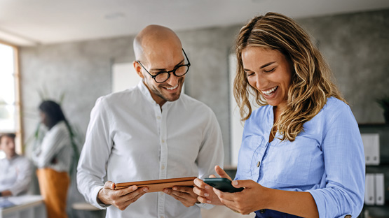 A woman in a blue shirt and a bald man in a white shirt, both smiling and looking at a smartphone held by the woman. The man is also holding a tablet. Other blurred colleagues are in the background.