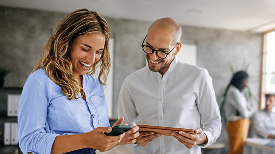 A woman in a blue shirt and a bald man in a white shirt, both smiling and looking at a smartphone held by the woman. The man is also holding a tablet. Other blurred colleagues are in the background.