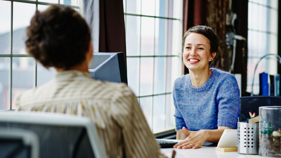 A smiling woman in her 40s with short brown hair wears a blue sweater and is talking to a coworker at her desk in an office with large windows.