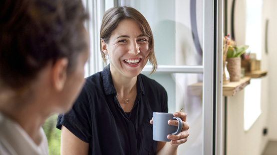 A person with short brown hair holds a mug while smiling and talking to someone inside an office.