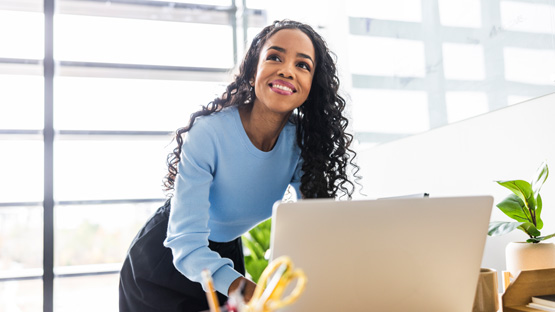 A smiling woman with long curly hair in her 30s, wearing a light blue shirt, uses a laptop at her desk near a bright window.