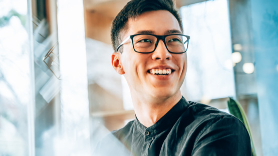 A smiling man in his 20s wearing glasses and a dark shirt looks happily to the side, with a blurry office scene reflected in a glass panel.