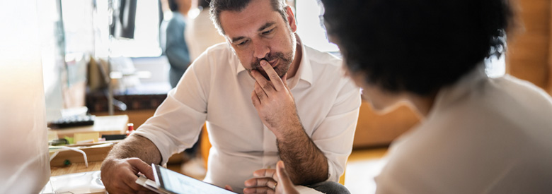 A person in a white collared shirt looks thoughtfully at a tablet held by another person while discussing something in a bright office.