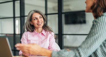 A smiling adult with gray hair in a pink shirt looks up and to the right while another person is blurred in the foreground.