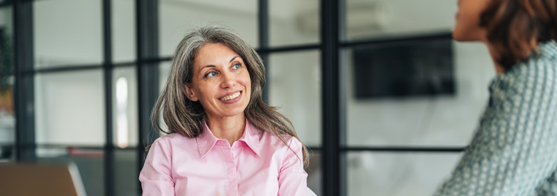 A smiling adult with gray hair in a pink shirt looks up and to the right while another person is blurred in the foreground.
