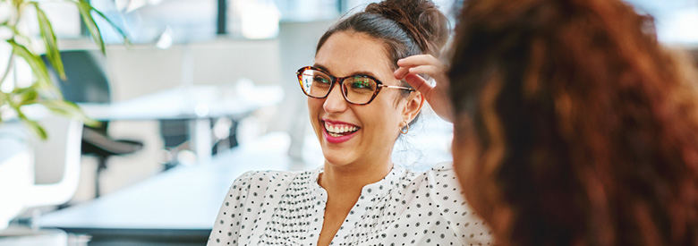A smiling woman wearing glasses and a polka-dotted shirt talks to another person.