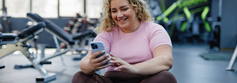 A young adult with curly blonde hair sits on the floor of a gym, smiling and looking at a phone.