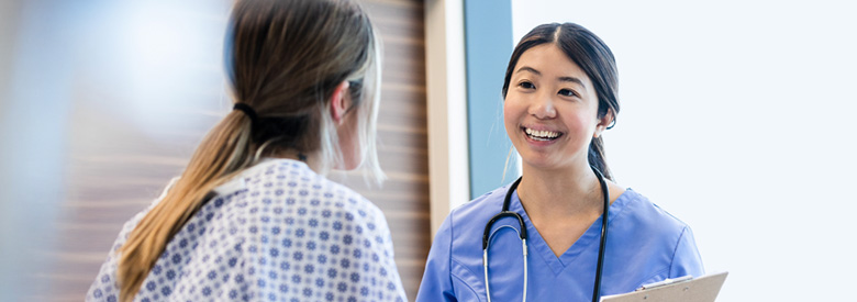 A smiling medical professional in a white lab coat and a medical professional in blue scrubs talk to a patient in a medical gown, offering comfort and support.