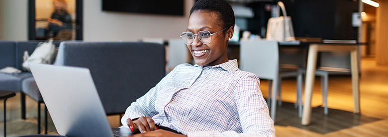 A smiling Black woman with short hair and glasses uses a laptop in a relaxed office setting, wearing a plaid shirt.