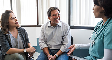 A smiling man and a woman in a grey cardigan listen attentively to a healthcare professional in turquoise scrubs.