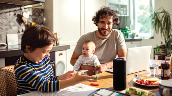 Man holding baby and smiling at young boy while sitting at a kitchen countertop listening to a Highmark health podcast through a speaker.