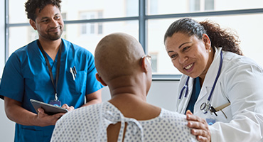 A medical professional in a white coat with a stethoscope and another medical professional in blue scrubs smile kindly at an adult patient in a hospital gown.