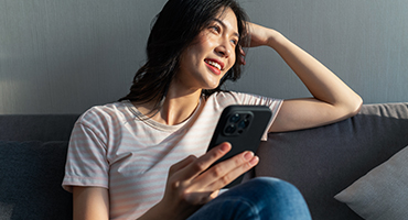 A young woman sits on a sofa, looking up and smiling while holding a smartphone in her hands. 