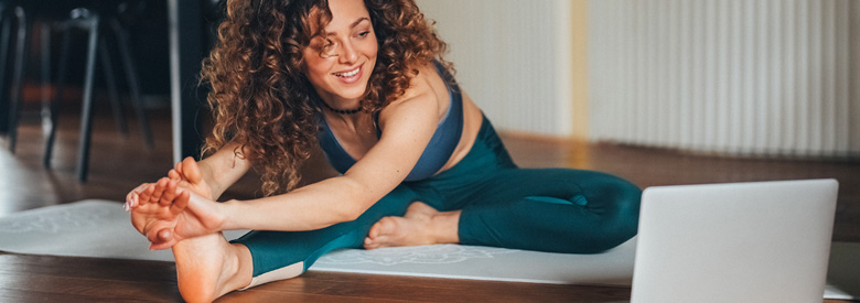Young adult with curly hair smiles while stretching during an online yoga class on a laptop.