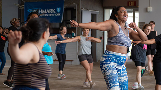 A group of women laughing and exercising in what appears to be a fitness class, with a Highmark Eastern New York banner visible in the background.