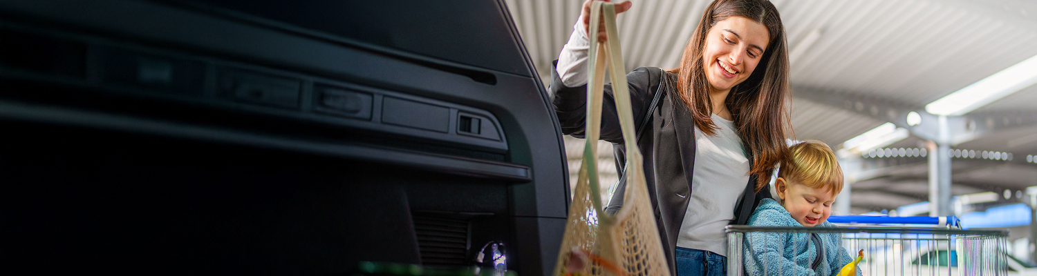 A happy young adult smiles while placing groceries into a car trunk, with a young child sitting in a shopping cart nearby.
