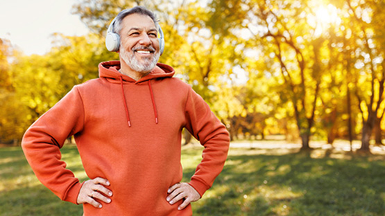An older man with a gray beard wearing headphones and an orange hoodie, standing outside in a park surrounded by autumn trees and warm sunlight.