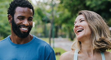 A man and woman laugh outdoors in a sunny setting.