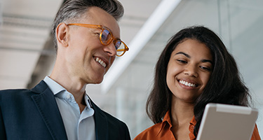 A smiling man with glasses looks towards a smiling woman holding a tablet.