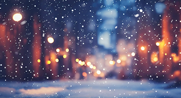 is a blurred, atmospheric shot of a city street at night during a snowfall. Soft, out-of-focus lights from streetlamps and buildings create warm glows against the cool, dark background.