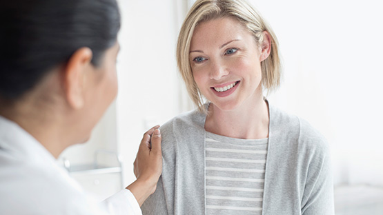 A medical professional in blue scrubs touches the shoulder of a smiling woman.