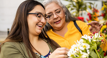 A young adult and an older adult are looking at flowers and smiling while at a flower shop.