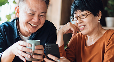 Two older adults are looking at a cell phone and smiling together, one holding a coffee mug.