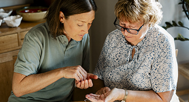 Two adults sit together at a table inside as one adult pours pills into the other's open palm.