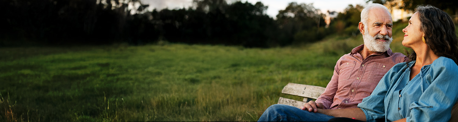 Two older adults sit outside on the grass and look at each other, smiling.