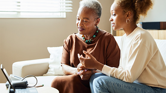 An older adult and a younger adult sit on a couch while looking at a laptop screen and discussing something important.