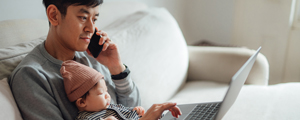 A parent is talking to a Highmark ACA expert on a mobile phone while holding a baby on their lap and working on a lap top computer.
