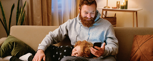 A man with a beard, wearing a gray hoodie, sits on a couch while looking at his phone. A child with red hair is lying with their head on his lap.
