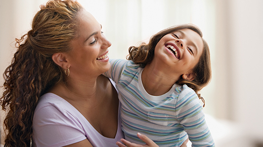 A woman with curly hair smiling and laughing happily while holding her young daughter, who is leaning back in joy.