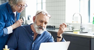 Grey-haired woman looks over shoulder of grey-bearded man sitting in kitchen with laptop looking at Highmark Medicare Plans