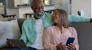 An older black couple on a couch, smiling and engaged with their smart devices, against a modern kitchen backdrop, illustrates "How Medicare Advantage Compares to Other Medicare Add-Ons."