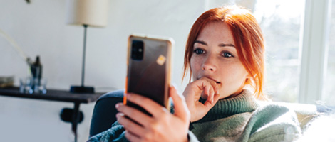 A young redhead woman looks thoughtfully at her phone, possibly worried or pondering something.